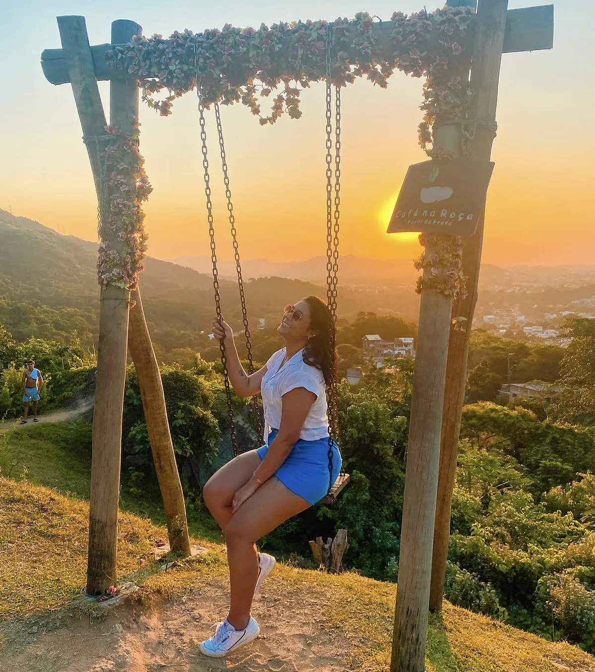 Mulher no balanço florido do Café na Roça, com vista para a mata e a cidade ao pôr do sol